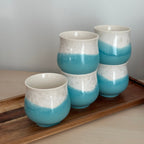 Set of four blue and white ceramic cups hanging on a wooden shelf against a light gray wall.
