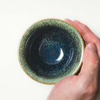Hand holding a ceramic tea cup with a textured green and black design on a white background