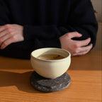 Ceramic cup with a dark liquid on a stone coaster, person sitting in the background